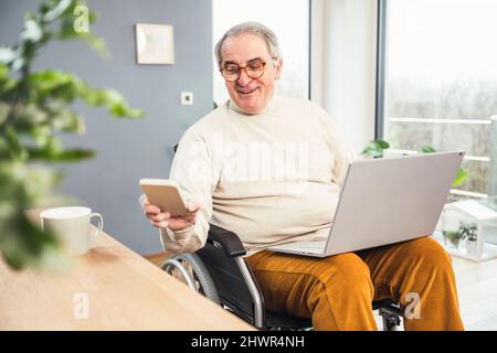 Smiling senior disabled man checking mobile phone sitting on wheelchair at home Stock Photo