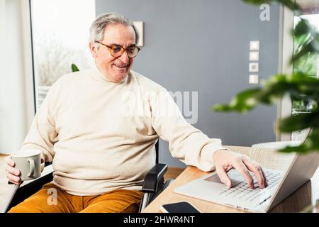 Smiling disabled man holding coffee cup using laptop sitting on wheelchair at table Stock Photo