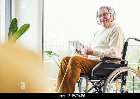 Smiling senior disabled man with tablet PC sitting on wheelchair at home Stock Photo