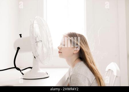 Girl sitting in front of table with electric fan at home Stock Photo