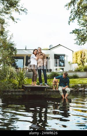 Man with woman standing by son and dog sitting by lake at backyard Stock Photo