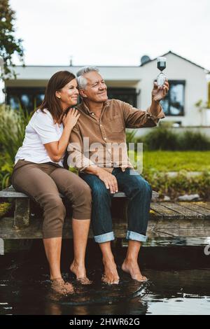 Smiling couple looking at hourglass sitting on jetty by lake Stock Photo