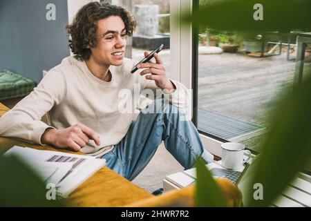 Smiling young man talking on smart phone through speaker in living room Stock Photo