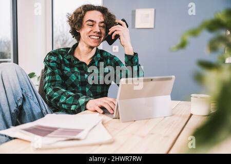 Happy young man talking on mobile phone through speaker sitting at table in living room Stock Photo