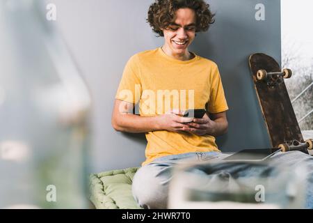 Happy man text messaging through smart phone by skateboard at home Stock Photo
