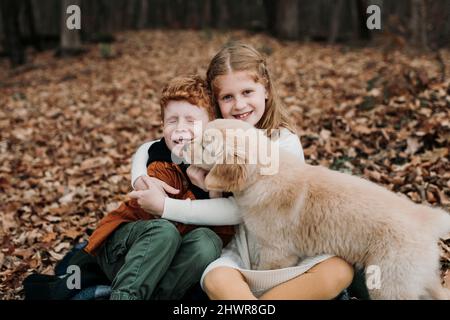 Puppy licking boy's face sitting with sister on autumn leaves in forest Stock Photo