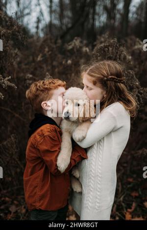 Brother and sister kissing golden retriever puppy in forest Stock Photo