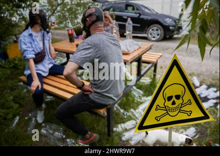 Selective focus of poison toxic sign in roadside forest with family and plastic bottles. Man, woman and child sitting at the table among garbage with skull-and-crossbones symbol on forefront. Stock Photo