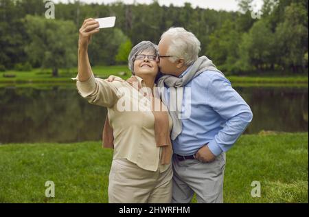 Cheerful old wife making selfie by phone camera while husband kissing her cheek Stock Photo