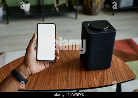 Background image of male hand holding phone with blank screen next to home AI smart speaker Stock Photo