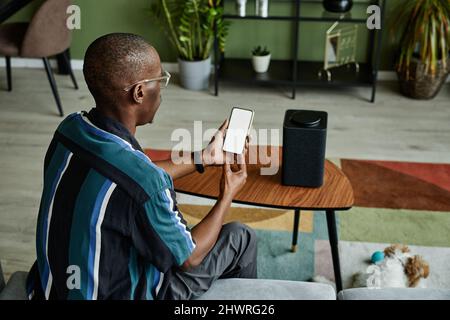 Back view view at modern black man connecting phone to smart speaker with voice controlled home AI Stock Photo
