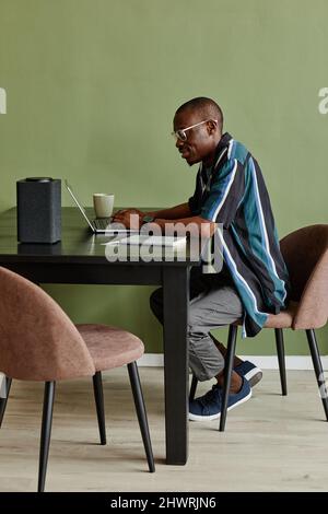 Vertical side view portrait African American man using laptop at home office with smart speaker against green wall Stock Photo