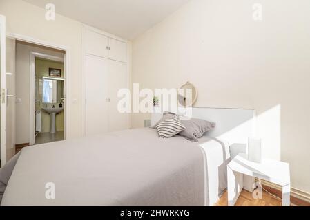 bedroom with king size bed with gray bedspread and cushions, white headboard, white wooden doors built-in wardrobe and en-suite toilet Stock Photo
