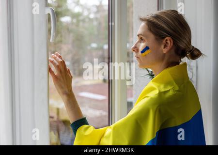woman support ukraine. wrapped in a flag looking out of window at home Stock Photo