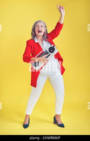 I bring the fun and noise wherever I go. Full length shot of a funky and stylish senior woman dancing while holding a boombox against a yellow Stock Photo
