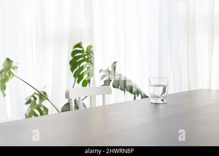 Glass of water placed on white table with chair near potted Monstera deliciosa plant in light modern apartment Stock Photo