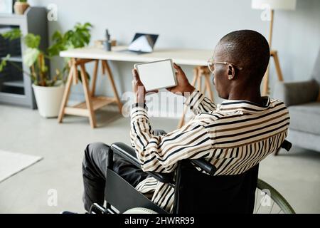 Back view of African American man in wheelchair using tablet with blank screen in minimal home interior Stock Photo
