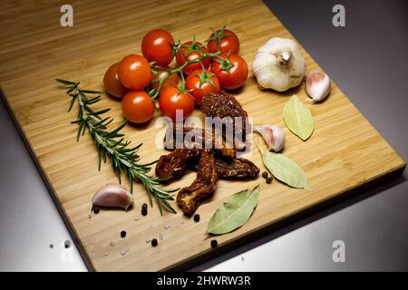 Vegetables and spices on a wooden board. Cherry tomatoes and sun-dried tomatoes, garlic, rosemary sprig, bay leaves, black pepper and coarse sea salt. Stock Photo