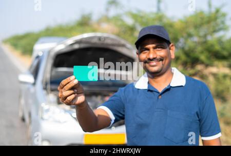 Smiling car mechanic showing green card by looking at camera in front of broken car - concept of promotion for mobile repair service. Stock Photo