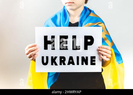 Boy wrapped with Ukrainian flag is holding a demonstration placard with Help Ukraine message. Russia vs Ukraine war Stock Photo