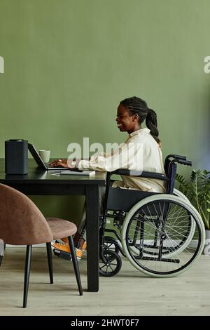 Vertical side view portrait of young African American woman using wheelchair while working at home office against green wall Stock Photo