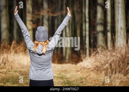 Woman exercising and stretching before running in forest. Happy female runner with arms raised. Warm up exercise during fitness activity Stock Photo