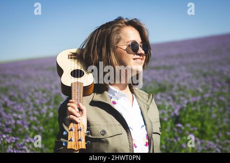 Happy young smiling woman with ukulele enjoying sunlight outdoors at spring Stock Photo