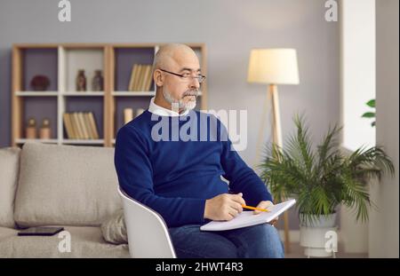 Serious writer sitting in a chair, holding a pencil and a notebook, looking away and thinking Stock Photo