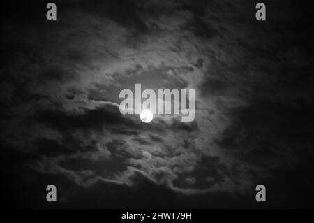 Full moon shining glowing light through the darkness of cloudy night sky. Black white photo Stock Photo