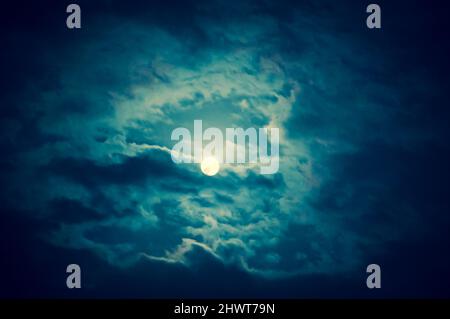 Full moon shining glowing light through the darkness of cloudy night sky. Moody blue photo Stock Photo