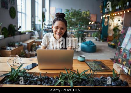 Joyous florist with cornrows sitting at a laptop in the workshop Stock Photo