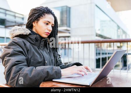 Side view of Hispanic female freelancer with Afro braids in warm jacket using laptop while sitting at table on terrace Stock Photo