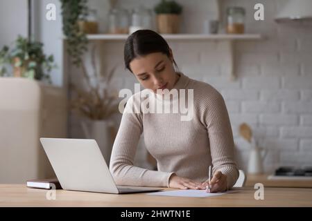 Woman sit at table with laptop jotting information on paper Stock Photo
