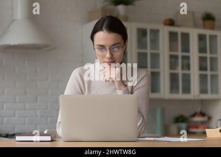 Pensive woman wear glasses use laptop seated in kitchen Stock Photo