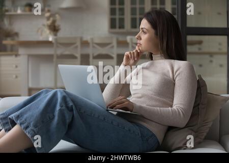 Thoughtful woman sit on couch with laptop staring away Stock Photo
