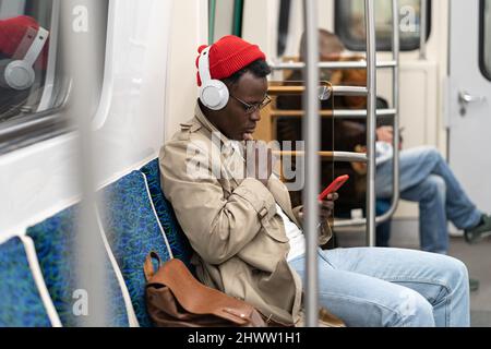 Pensive African young man reading posts, listening music, using smartphone, riding home by subway. Stock Photo