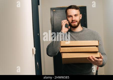 Bearded delivery man standing by door of customer apartment with boxes of hot pizza and calling customer on phone, looking at camera. Stock Photo