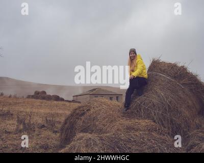 Laughing woman sits on rolled haystacks in abandoned area among the hills covered with cloudy haze Stock Photo
