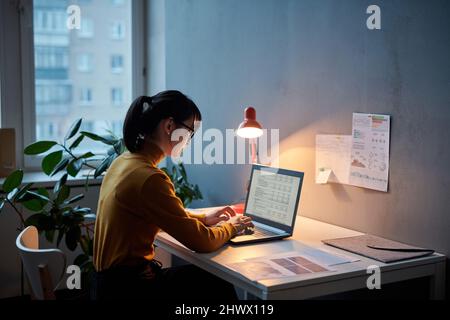 Young businesswoman sitting at her workplace and working with project online using laptop, she working till late at office Stock Photo