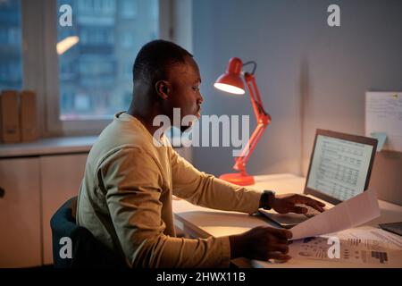 African businessman working over project till late evening at office, he sitting at table and typing graphs on laptop Stock Photo