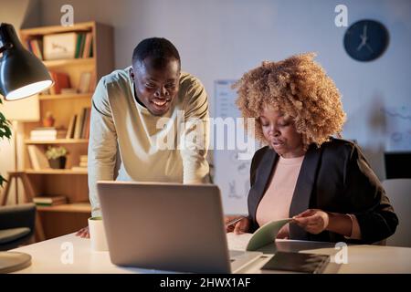 African woman examining the project sitting at table in front of laptop and discussing it with her colleague, they working in team till late night at Stock Photo