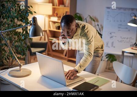African young businessman using laptop at table while talking on mobile phone with colleague, he working till late at office Stock Photo