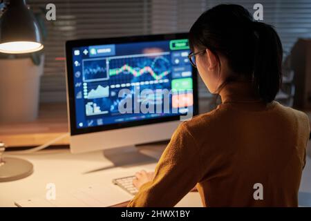 Rear view of young computer engineer sitting at table in front of computer monitor and developing new software working till late night at office Stock Photo