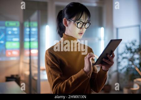 Asian young businesswoman in eyeglasses using digital tablet to work online while standing at office Stock Photo