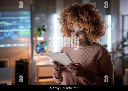 African young woman with curly hair watching something on digital tablet while working at office till late evening Stock Photo