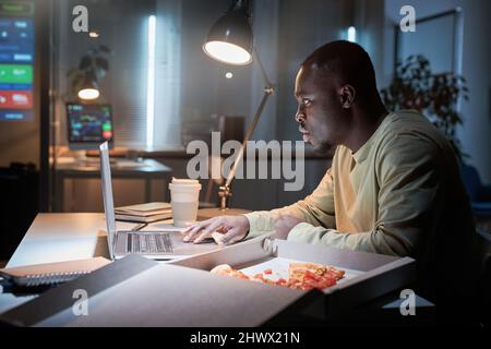 African businessman sitting at workplace and having a snack with pizza and coffee while working on laptop at office till late night Stock Photo