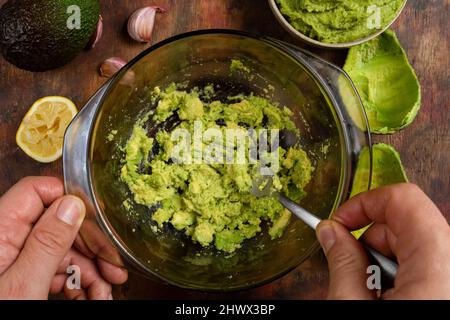 Man's hands mashing several avocados in a bowl with a fork to prepare a rich and fresh guacamole. Ingredients in composition on a wooden background in Stock Photo