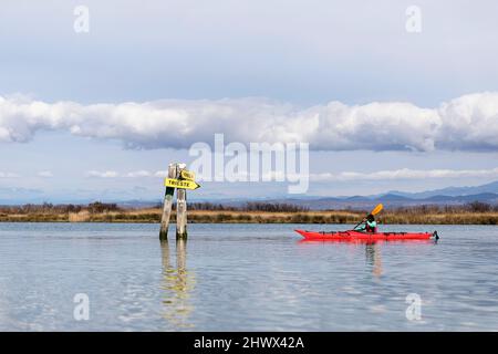 Woman by sea kayak exploring Isonzo river's mouth (Soča river) and stopping by direction signs - Regional Nature Reserve of the Foce dell'Isonzo,Italy Stock Photo