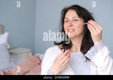 Woman touching clean side eye contour with an anti-bagging pipette, serum essence oil, looking away wearing bathrobe while sitting in bedroom on bed. Concept wrinkle care products in wrinkle at home Stock Photo