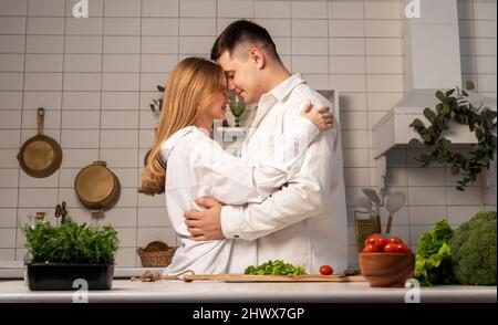 Happy young couple hugging while cooking at home. Handsome man and beautiful woman talking and smiling in modern scandinavian style kitchen. Healthy food concept. High quality photo Stock Photo